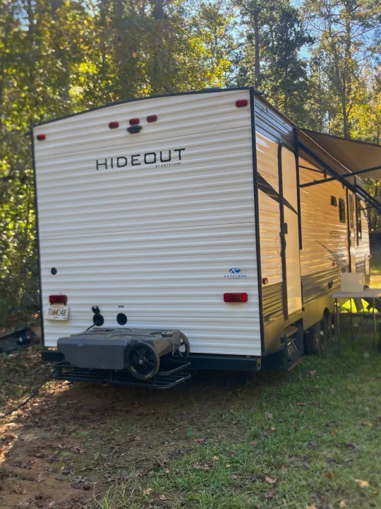 Keystone Hideout RV rental in a wooded setting, showing the rear with a waste tank on a rack and side awning.. Keystone RV Hideout 2021