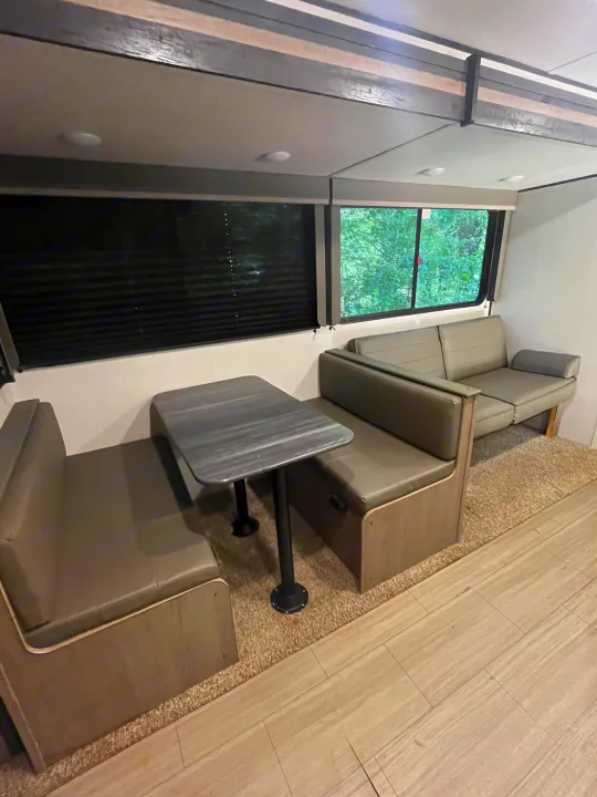 Interior of an RV rental showing a neutral-toned dining booth with a table, a sofa, and windows with outdoor views.. Keystone RV Hideout 2021
