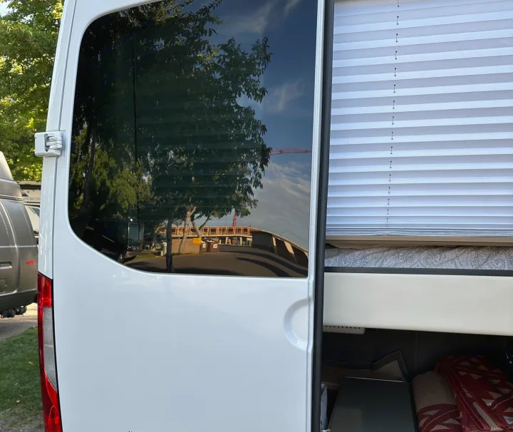 White RV rental with tinted rear door window reflecting trees, road, and construction. Interior shows pleated blind and bed.. Coachmen Galleria 2020