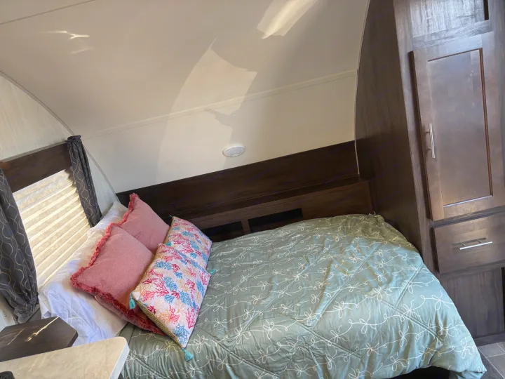 An inviting RV rental bedroom featuring a bed with a green patterned comforter, vibrant pillows, and wooden storage.. Forest River Cherokee 2019