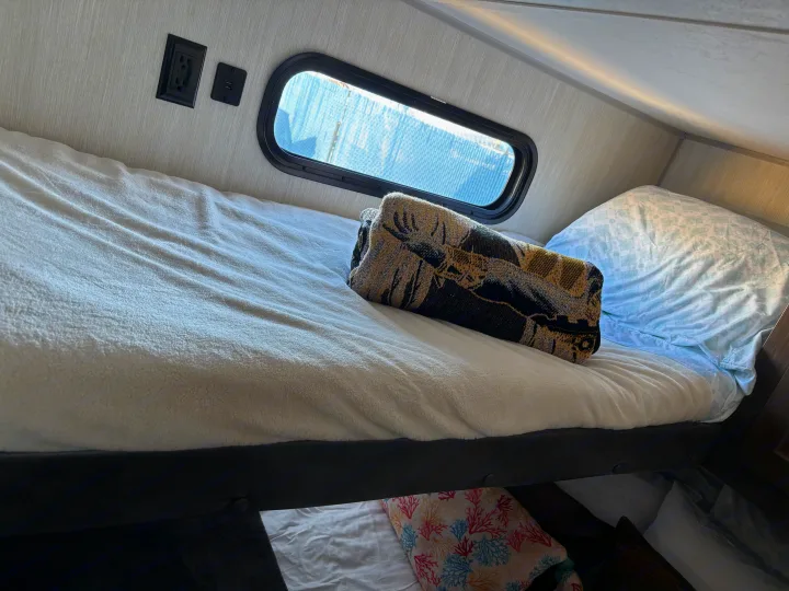 Inside an RV rental, a cozy bunk bed features a window, charging ports, and warm bedding, ideal for travel accommodation.. Forest River Cherokee 2019