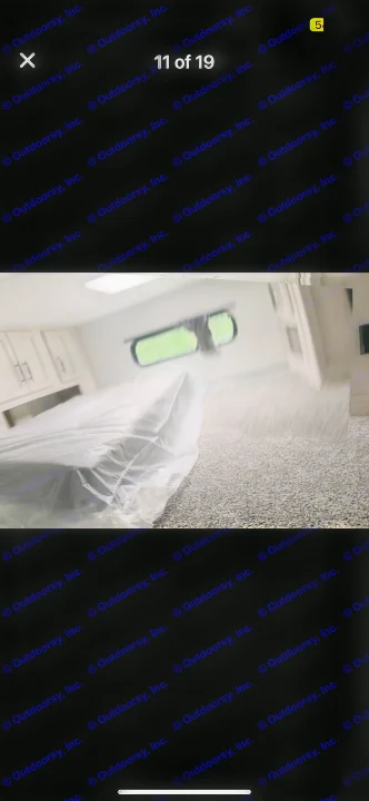 RV rental interior: bedroom with new plastic-wrapped bed, cabinets, and a window overlooking greenery. Gray carpeted floor.. Forest River Sandpiper 2024