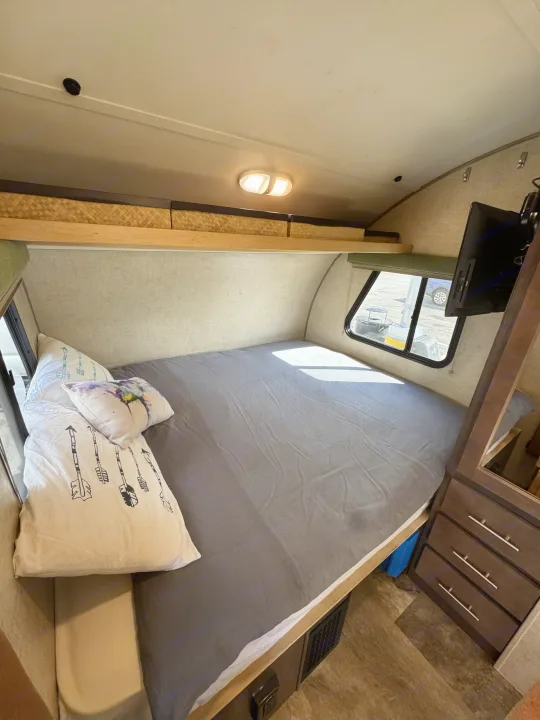 Interior view of an RV rental bedroom with a full bed, gray sheets, patterned pillows, overhead storage, wall-mounted TV, and window.. R-Pod Hood River Edition 2015