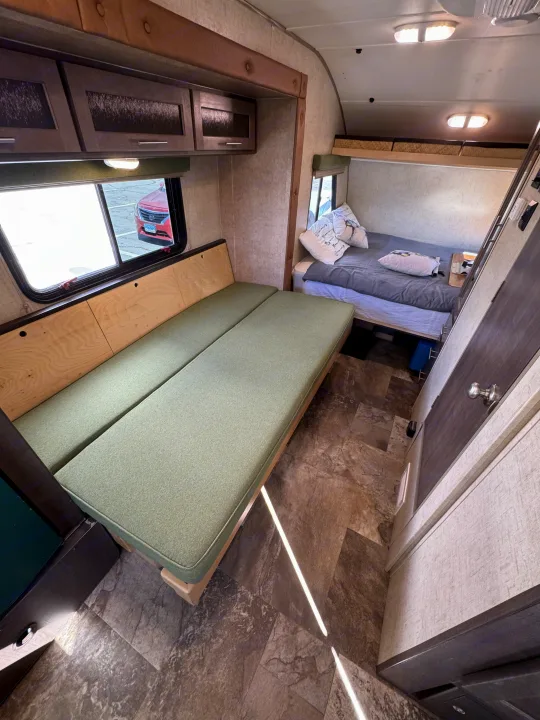 Interior view of an RV rental: green bench seating, overhead cabinets, a cozy bed, and a window view.. R-Pod Hood River Edition 2015