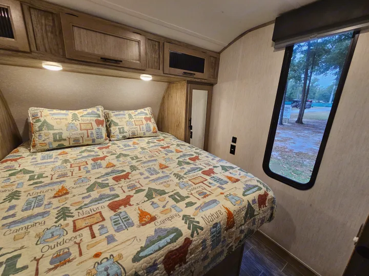 Inside an RV rental: a cozy bedroom with a camping quilt, storage cabinets above, and a window overlooking nature.. Forest River Cherokee Alpha Wolf 2021