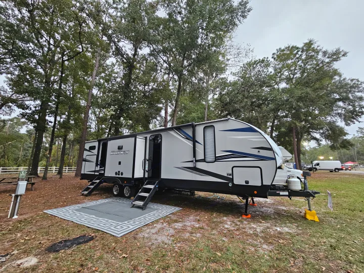 Grey and black rv rental travel trailer parked at a wooded campsite, featuring open steps and an outdoor rug.. Forest River Cherokee Alpha Wolf 2021