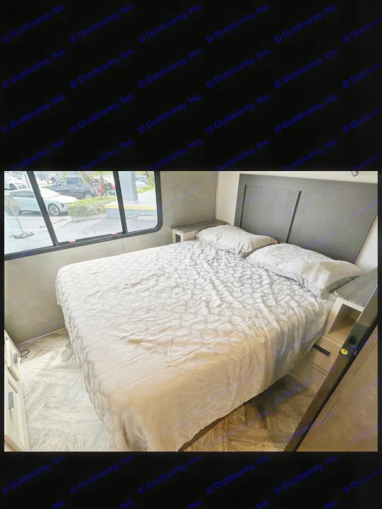 Cozy RV rental bedroom featuring a queen bed with geometric bedding, dark headboard, nightstands, and window showing parking lot.. Forester LE 2023