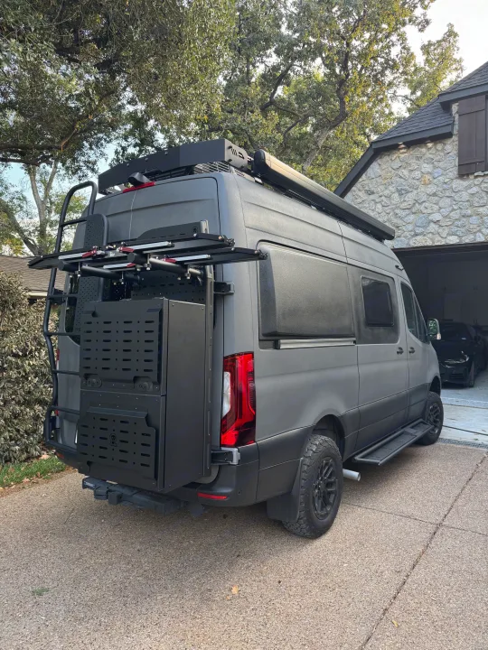 Customized Mercedes Sprinter RV rental: A rugged, dark grey van with off-road tires, roof rack & rear storage, parked on a driveway.. Winnebago Revel 2025