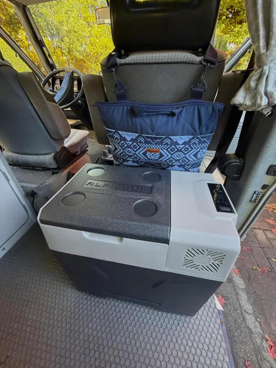 Inside an RV rental: Portable Alpicool fridge and Columbia bag are ready for adventure. VW steering wheel & autumn trees outside.. Volkswagen T3 Westfalia 1991