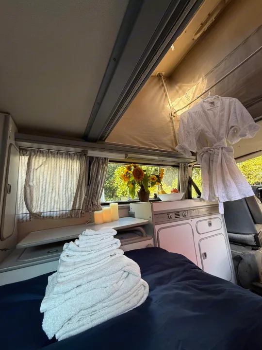 Cozy rv rental interior featuring a bed with fresh towels, sunflowers, and a kitchenette, perfect for a comfortable road trip.. Volkswagen T3 Westfalia 1991