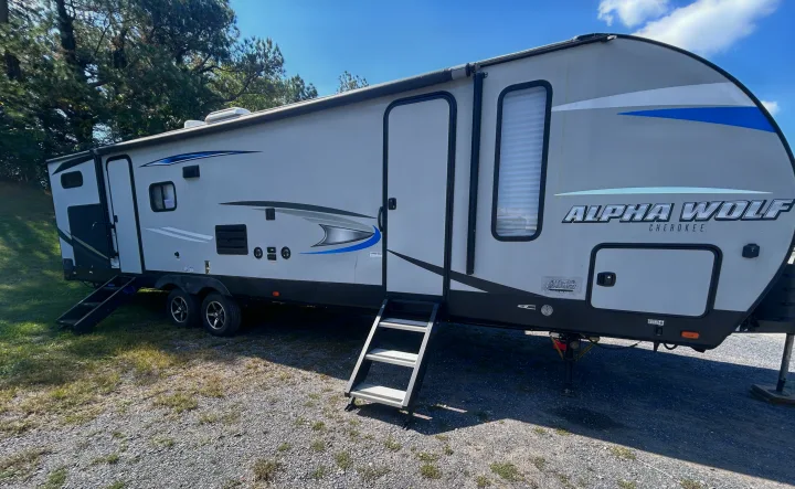 Gray and blue Alpha Wolf Cherokee RV rental parked on gravel with stairs leading to its doors, set against a clear sky and trees.. Escape Trailer Alpha Wolf 2020