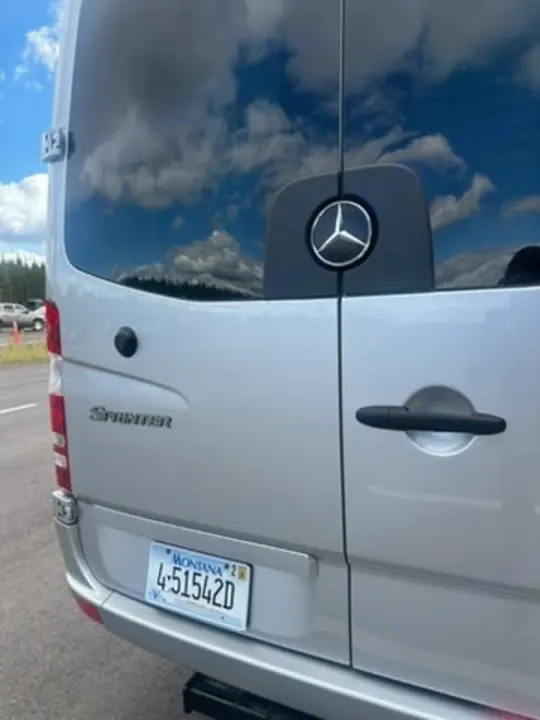 Rear view of a silver Mercedes Sprinter rv rental van, Montana plate visible. Blue sky & clouds reflect in its windows.. Coachmen Galleria 2020