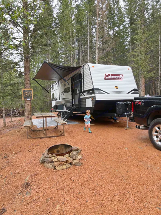 RV rental and truck at campsite #20, with child, picnic table, fire pit, surrounded by tall trees.. Dutchmen Coleman Lantern 2022