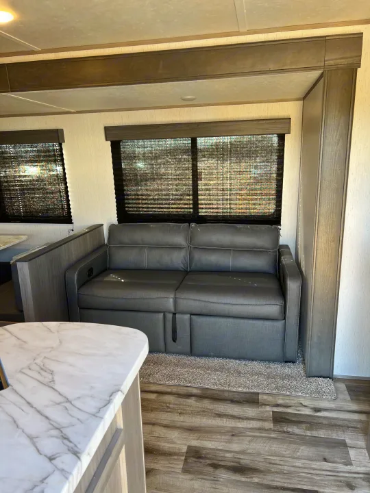 This modern RV rental living space features a gray pull-out sofa, large windows with blinds, wood-look floors, and a marble-pattern counter.. Dutchmen Coleman Lantern 2022