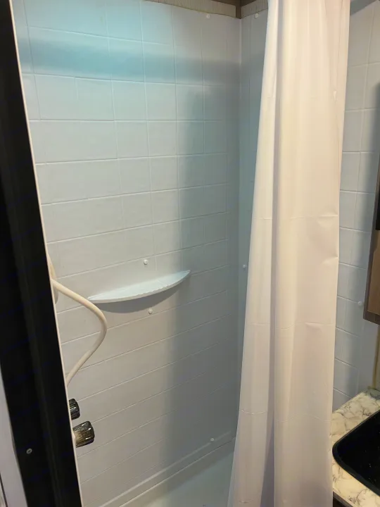 RV rental bathroom interior: A clean, compact shower stall featuring white tile-patterned walls, a shelf, and a shower curtain.. Dutchmen Coleman Lantern 2022