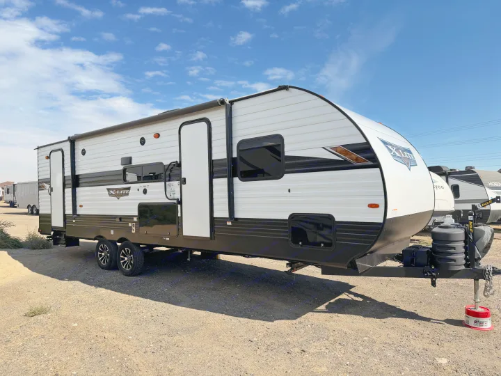 This spacious white & black travel trailer, ideal for rv rental, is parked on a gravel lot beneath a clear blue sky.. Forest River Wildwood X-Lite Platinum 2024
