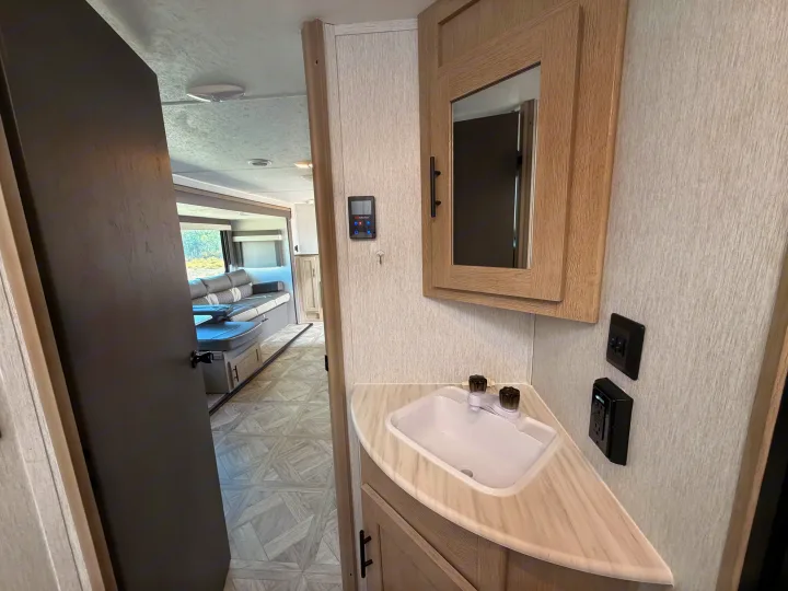 An RV rental's bathroom features a corner sink, mirrored cabinet, and controls, with a glimpse of the living space beyond.. Forest River Wildwood X-Lite Platinum 2024