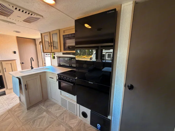 This sleek RV rental kitchen features a black Furrion fridge, stove, microwave, sink, and light wood cabinetry.. Forest River Wildwood X-Lite Platinum 2024