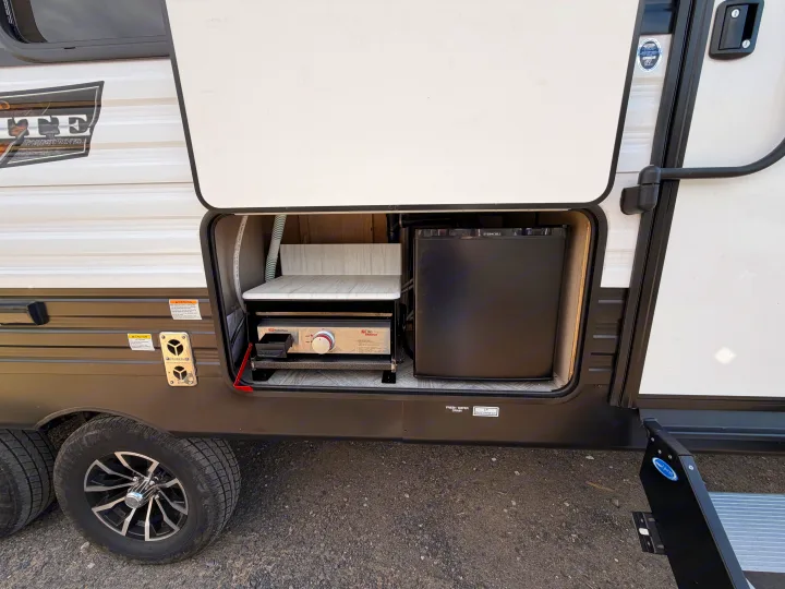 RV rental's exterior shows an open outdoor kitchen with a Suburban griddle, Everchill mini-fridge, and Step Above stairs.. Forest River Wildwood X-Lite Platinum 2024