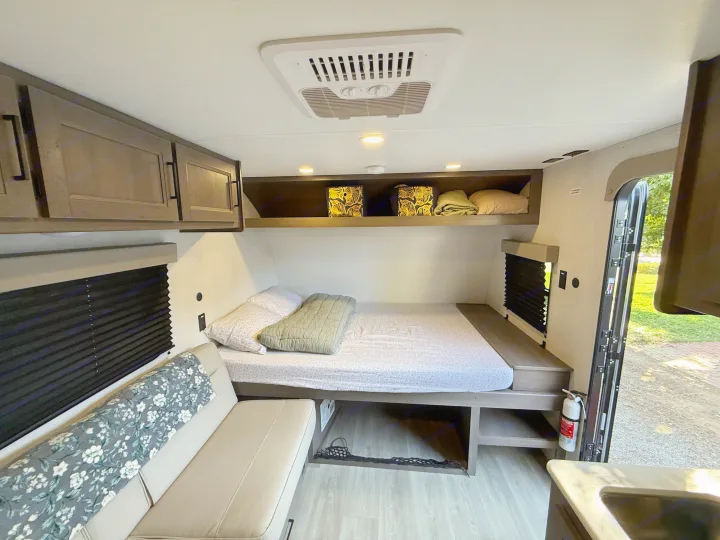This cozy rv rental interior features a bed on the right, a couch on the left, overhead cabinets, and an AC unit on the ceiling.. Grand Design Transcend One 2025