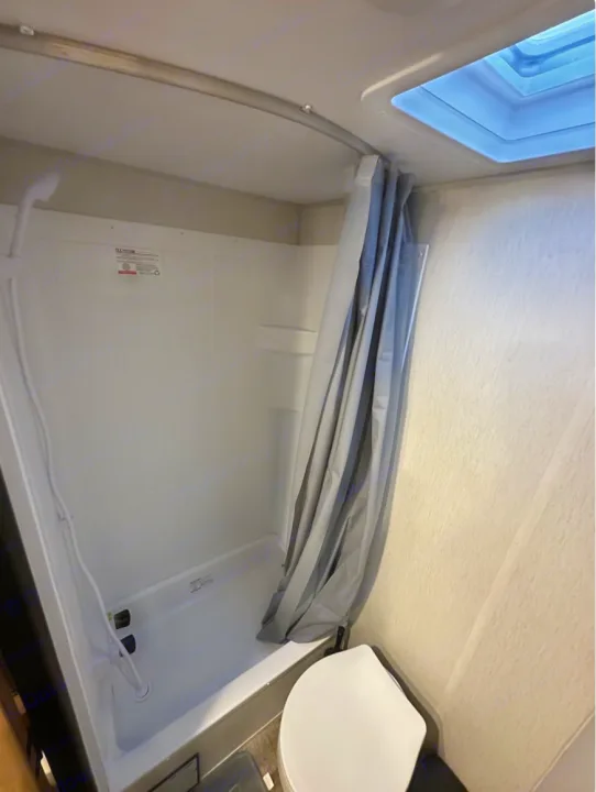 Compact RV rental bathroom featuring a white shower with a handheld showerhead, a gray curtain, and a skylight.. Forest River Cherokee Wolf Den 16 EV 2024