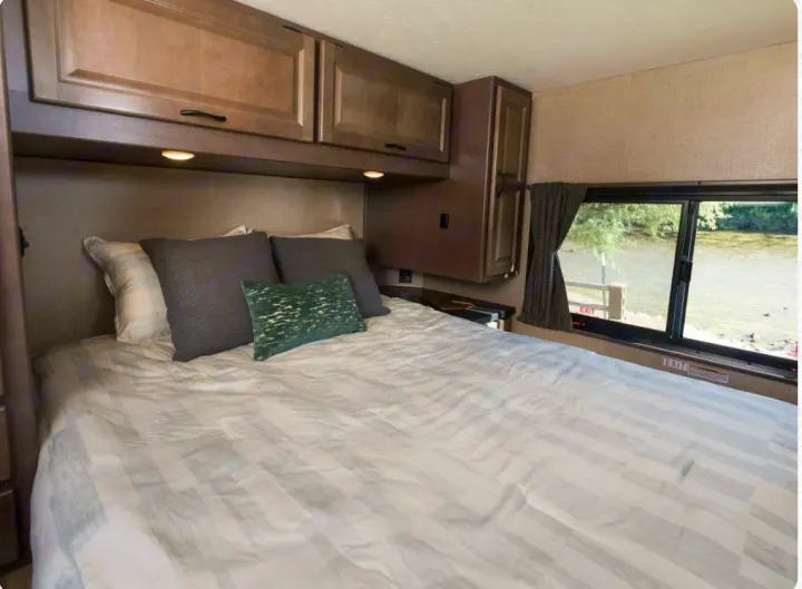 Cozy RV rental bedroom featuring a comfy bed, overhead cabinets, and a scenic window view overlooking water.. Thor Majestic 2019