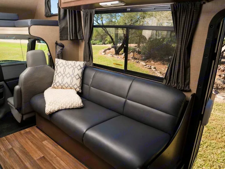 RV rental interior: comfy black sofa, pillows, and a large window with a scenic outdoor view for a relaxing journey.. Thor Majestic 2019