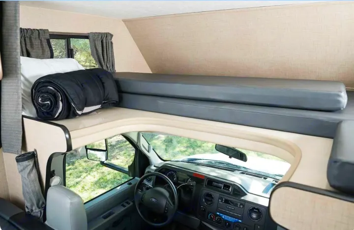 Inside an RV rental: view of the driver's cockpit below a cab-over bunk with a pillow and rolled comforter, ready for rest.. Thor Majestic 2019