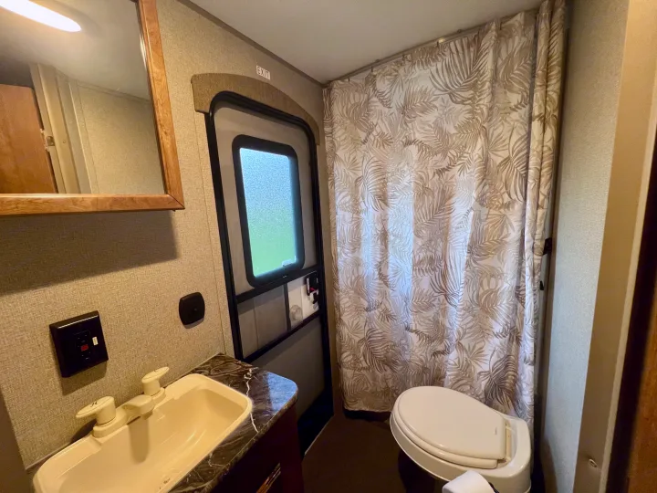 Compact RV rental bathroom interior features a sink, mirror, toilet, and leaf-patterned shower curtain, beside an exit door.. Keystone Springdale 2017