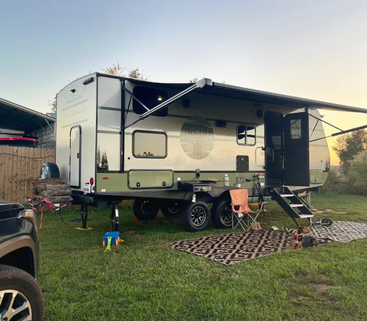 A modern RV rental, a Forest River NoBo travel trailer, is parked at a grassy campsite with an extended awning, outdoor gear, and a relaxed dog.. Forest River 5NHTRDV27TD211453 2026