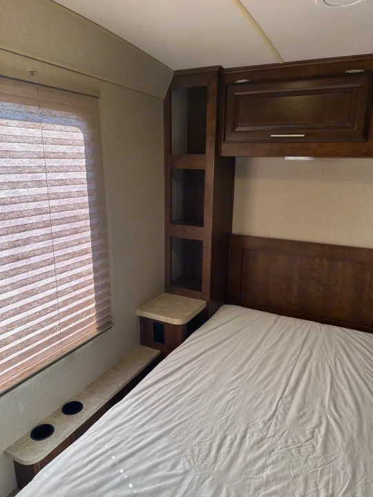 Comfortable RV rental bedroom with a bed, dark wood built-in storage, window blinds, and cup holders on a side shelf.. Forest River FR3 2017