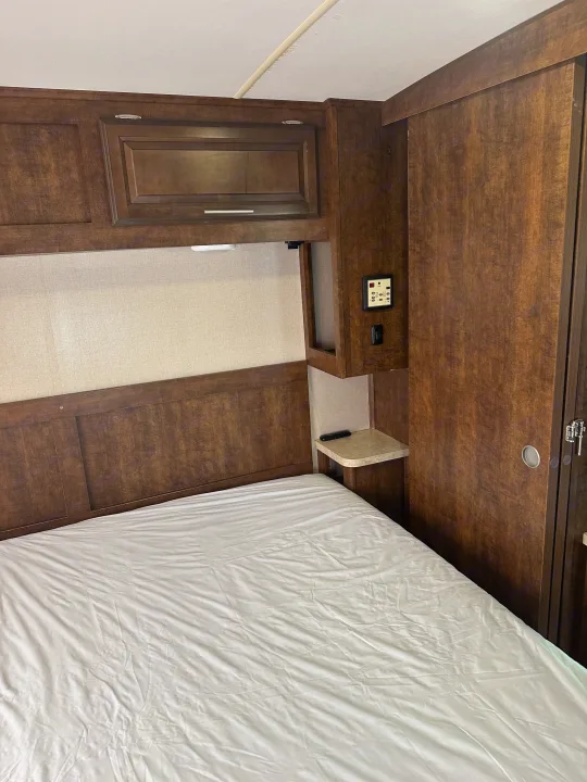 Cozy RV rental bedroom with a white-sheeted bed, dark wood cabinetry, and bedside controls. Perfect for your next adventure.. Forest River FR3 2017