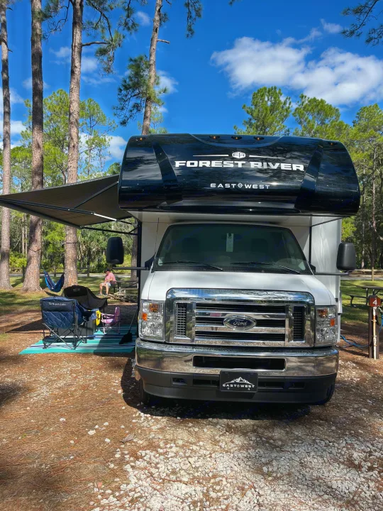 Perfect campsite scene with an RV rental: a white Forest River RV, awning out, chairs, hammock, and tall pines under a blue sky.. East to West Entrada 2025