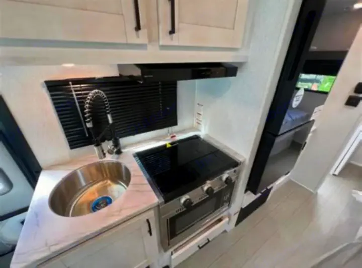 Bright RV rental kitchen featuring a stainless sink, 2-burner stove, oven, and marble-look counter with light cabinets.. East to West Entrada 2025
