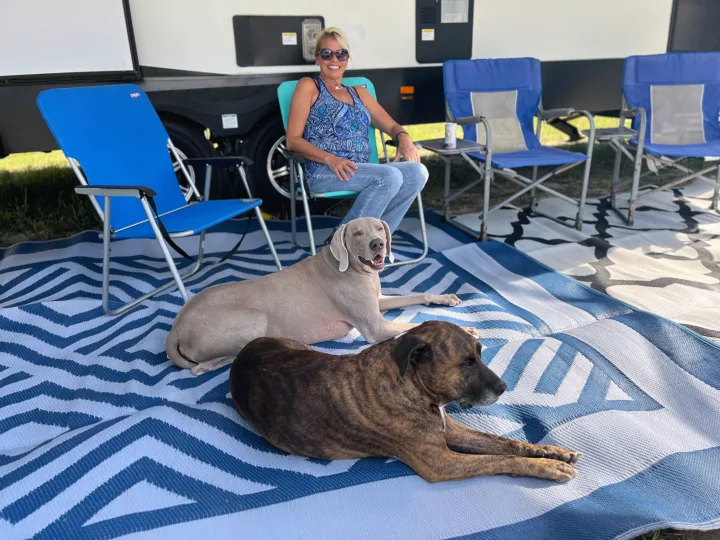 RV rental fun! A woman and her two dogs relax on a blue and white mat with camping chairs beside their RV.. East to West Alta 2022