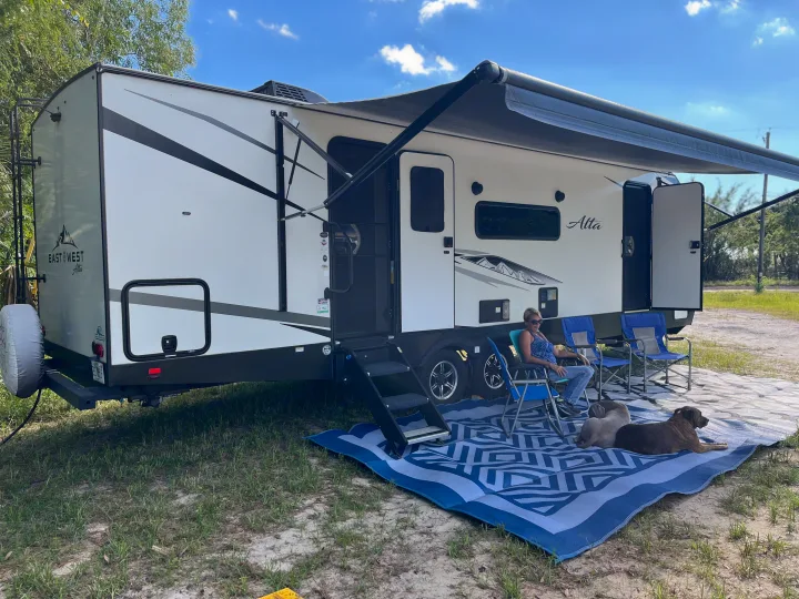 A person and two dogs relax outside a white Alta RV rental, enjoying the campsite under the extended awning on a blue mat.. East to West Alta 2022