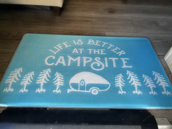 Light blue "Life is Better at the Campsite" mat with trees & camper graphic inside an RV rental.. East to West Alta 2022