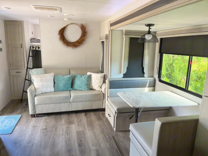 RV rental interior with a light sofa, throw pillows, decorative wreath, dinette, and window views of lush greenery.. East to West Alta 2022