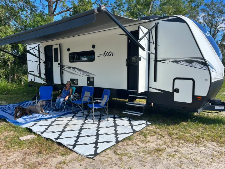 Serene RV rental camping scene: a woman and two dogs relax on outdoor mats under the awning of their travel trailer.. East to West Alta 2022