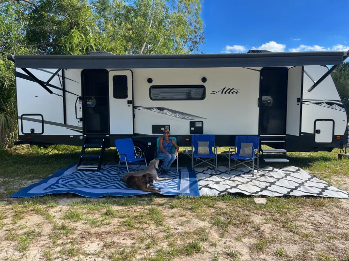 Enjoying an RV rental adventure: a woman and two dogs relax outside their camper with chairs and rugs at a sunny campsite.. East to West Alta 2022
