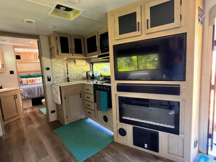Modern RV rental interior with light-toned kitchen, entertainment center with TV/fireplace, and a cozy bedroom view.. East to West Alta 2022