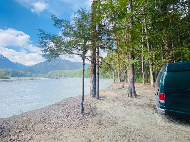 RV rental van, dark green, parked by a wide, glacial river & mossy forest. Snowy mountains under blue sky.. Ford Econolline 250s 2001
