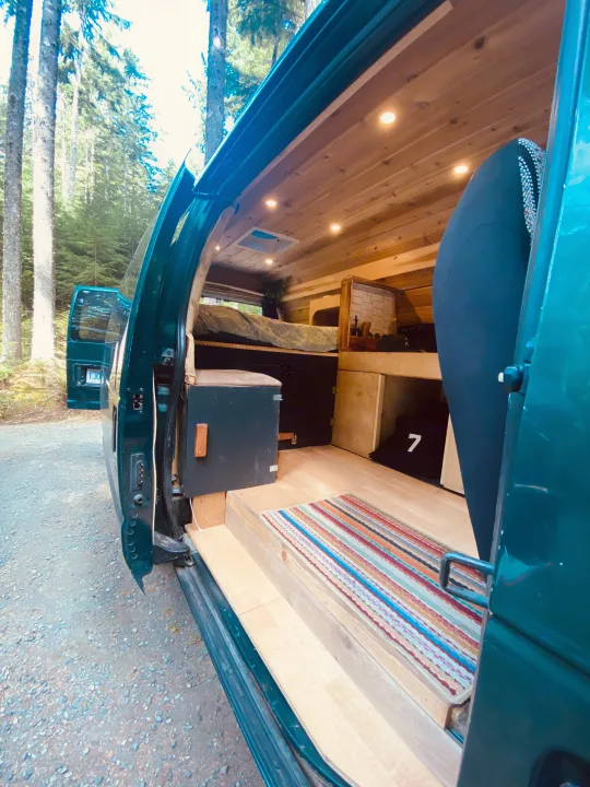 Converted RV rental van interior: a cozy wood-paneled space with bed, storage, and rug, amidst tall forest trees.. Ford Econolline 250s 2001