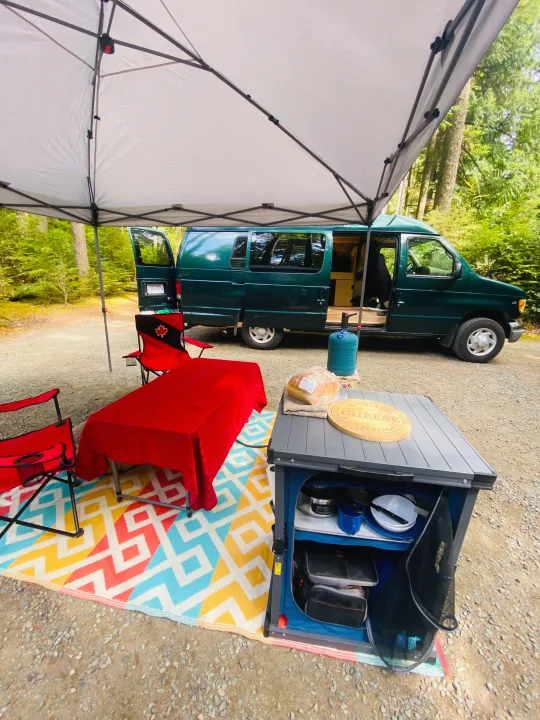 Cozy RV rental camping setup in the forest with an outdoor kitchen, table, and chairs next to a dark green van.. Ford Econolline 250s 2001