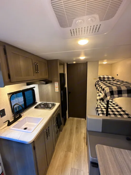 This RV rental interior features a compact kitchen, bunk beds with plaid bedding, a dinette, and wood-look flooring.. Keystone RV Hideout Sport 2025