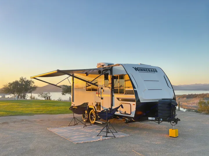 Scenic RV rental setup: Winnebago travel trailer by a lake at sunset, with chairs and a rug laid out.. Winnebago Micro Minnie 2100BH 2025