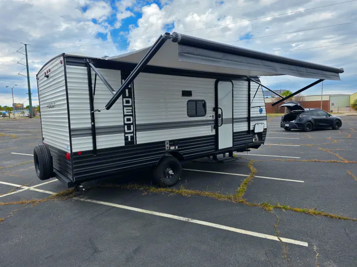 An RV rental, a white travel trailer with its black awning extended, is parked in a paved lot beside a black car with its trunk open.. Keystone RV Hideout Sport 2025