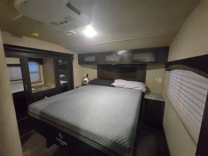 Inside an RV rental: a cozy bedroom features a Sapphire Sleep mattress, cabinets, fan, and window blinds.. Forest River Shockwave 2018