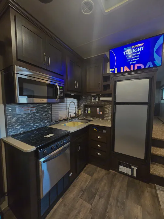 Spacious RV rental kitchen with dark wood cabinets, stainless steel appliances, modern backsplash, and TV mounted above fridge.. Forest River Shockwave 2018