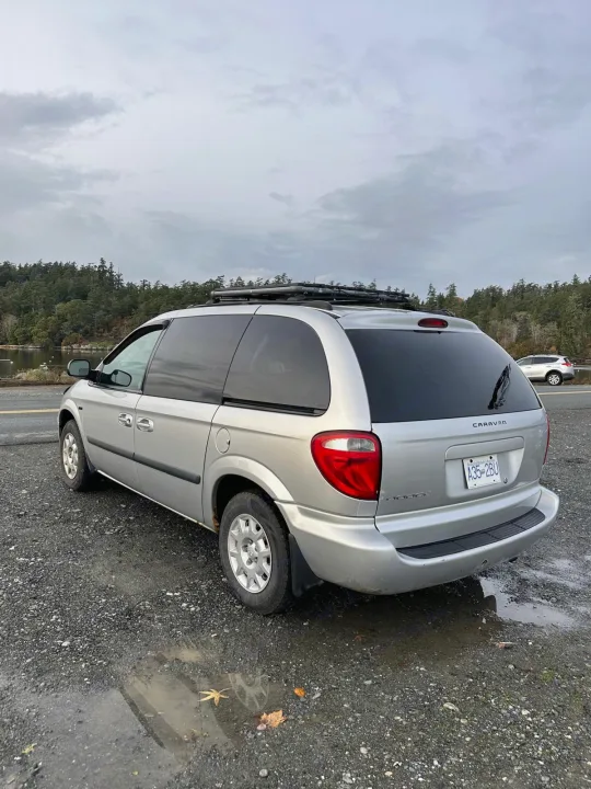 Silver Dodge Caravan minivan, ideal for an rv rental road trip, parked by a scenic lake with roof rack under cloudy skies.. Dodge Caravan 2005